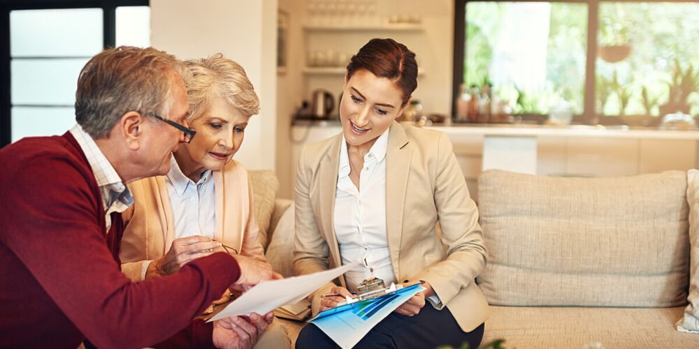 Angehörigen sitzen auf einem Sofa und schauen sich gemeinsam mit einer Pflegeberaterin Dokumente an. Die Beraterin lächelt, während sie mit dem Paar spricht. Im Hintergrund sind Pflanzen und eine helle, moderne Küche zu sehen.