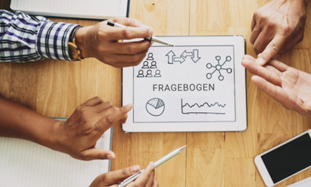 Eine Gruppe von Personen diskutiert über einen Fragebogen auf einem Tablet. Der Bildschirm zeigt das Wort "FRAGEBOGEN" sowie verschiedene Symbole, die sich auf Bedarfsanalysen im Pflegebereich beziehen. Die Hände der Teilnehmer sind sichtbar, während sie