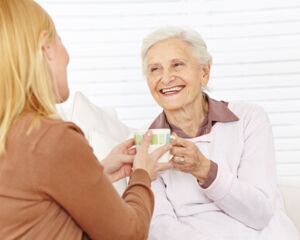 Eine Betreuerin mit einer brauen Bluse sitzt mit einer älteren Dame mit hellem Oberteil und grauen Haaren auf einem Sofa und reicht ihr eine Tasse. Beide lächeln und scheinen ein angenehmes Gespräch zu führen. Die Umgebung ist hell und freundlich.