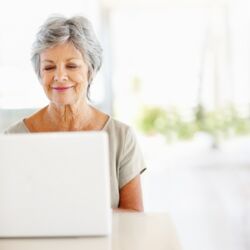 Eine ältere Frau sitzt an einem Tisch lächelnd auf einen Laptop. Die Umgebung ist hell und freundlich, mit unscharfen Pflanzen im Hintergrund. Das Bild vermittelt einen Eindruck von Pflegeberatung und digitaler Unterstützung im Zusammenhang mit Pflegegrad