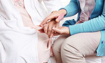 Eine Hand von einer Betreuerin hält die Hand einer älteren Dame, während sie auf einem Bett sitzen. Die Betreuerin trägt einen blauen Pullover und zeigt eine fürsorgliche Geste. Die ältere Person trägt einen weißen Bademantel und einen Schlafanzug.