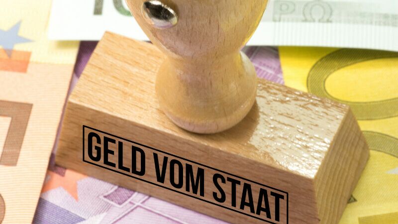 Holzstempel mit der Aufschrift "GELD VOM STAAT", der auf Geldscheinen platziert ist. Der Stempel deutet darauf hin, dass es sich um staatliche finanzielle Unterstützung oder Mittel handelt.