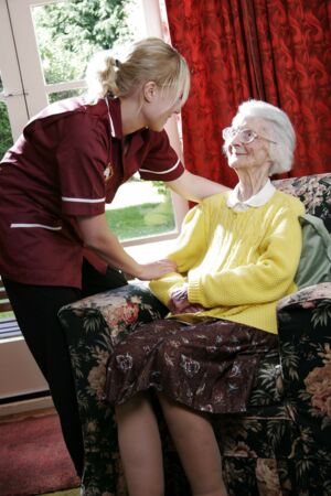 Eine Pflegekraft spricht freundlich mit einer älteren Dame, die in einem gemütlichen Sessel sitzt. Die Pflegekraft trägt eine uniformähnliche Kleidung und zeigt Fürsorge, während die ältere Dame in einem gelben Pullover lächelt