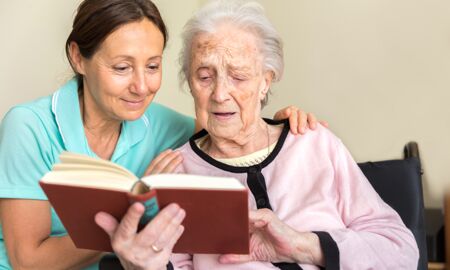 Eine Betreuerin liest gemeinsam mit einer älteren Frau in einem Pflegeheim aus einem Buch. Die Betreuerin lächelt und zeigt Interesse, während die ältere Frau aufmerksam zuhört und das Buch betrachtet.