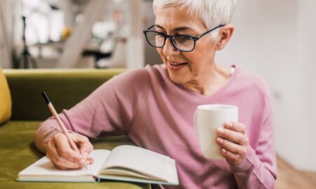 Eine Dame mit kurzen, grauen Haaren sitzt auf einer grünen Couch und schreibt in ein Notizbuch. Sie hält eine Tasse in der Hand. Sie trägt einen rosa Pullover und wirkt konzentriert und zufrieden. Das Bild vermittelt ein Gefühl von Ruhe in der Pflege