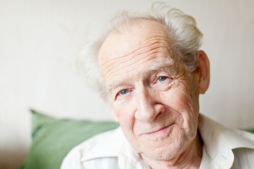 Ein älterer Mann mit grauen Haaren und einem sanften Lächeln sitzt auf einem grünen Kissen. Er hat eine nachdenkliche Ausstrahlung, die die Bedeutung von Altenpflege widerspiegelt