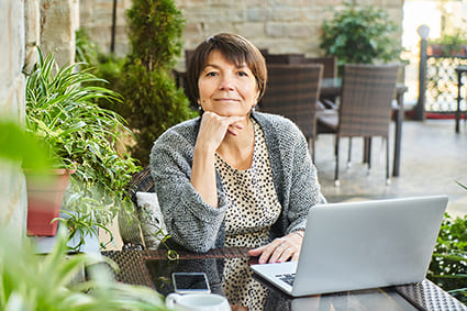 Eine Frau sitzt an einem Tisch im Freien mit einem Laptop und einem Smartphone, umgeben von Pflanzen. Sie hat einen nachdenklichen Ausdruck und trägt einen grauen Pullover. Das Bild vermittelt eine entspannte Atmosphäre mit dem Thema Pflege